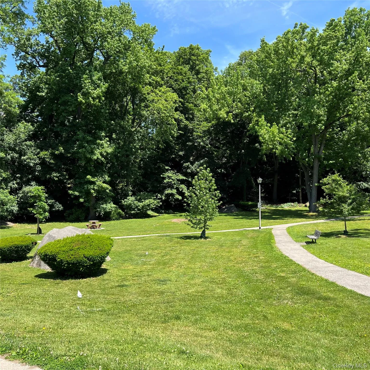 Expansive green space featuring a paved walking path, mature trees, manicured hedges, and a picnic tables Expansive green space featuring a paved walking path, mature trees, manicured hedges, and a picnic tables