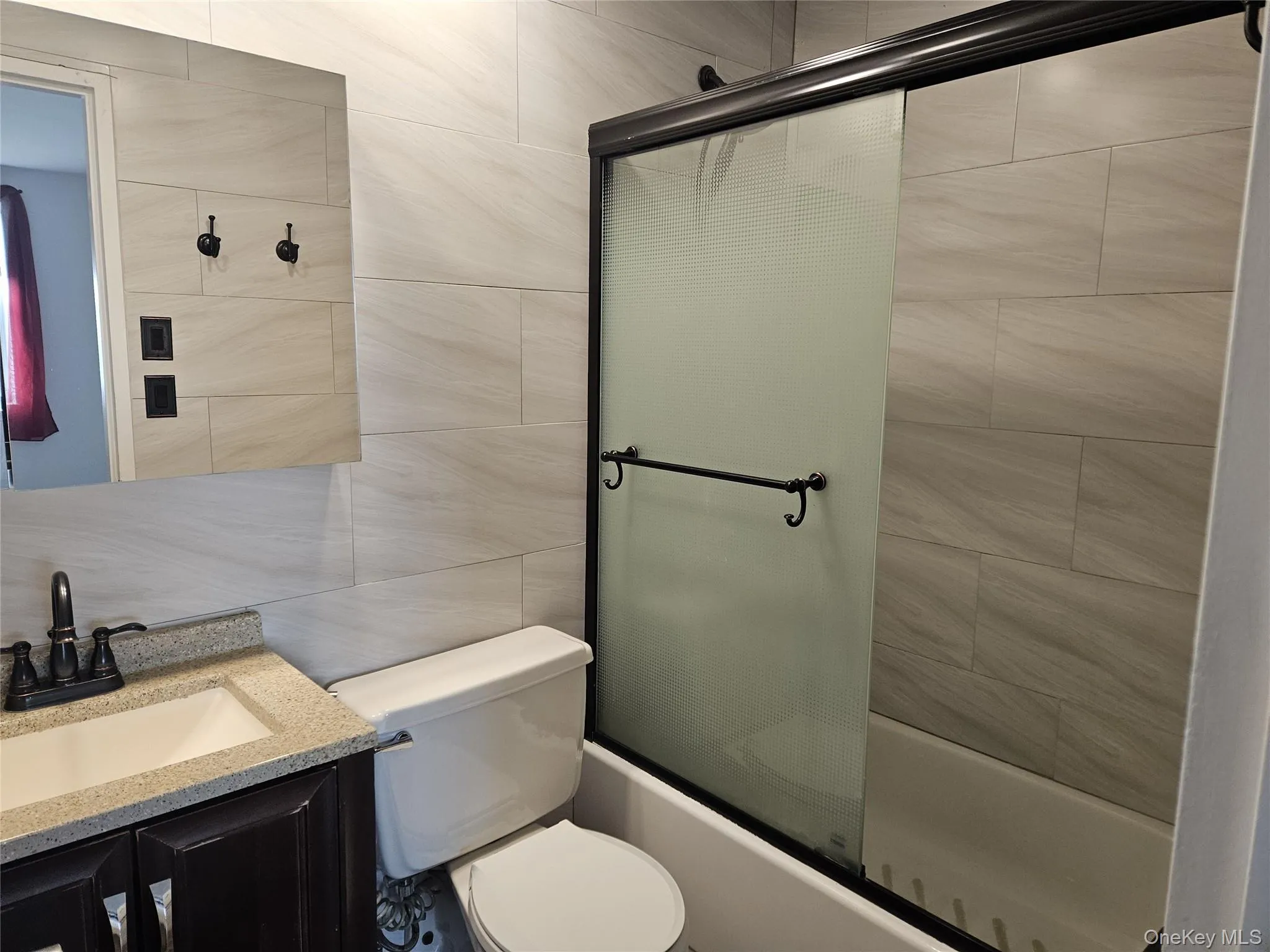 Full bathroom with vanity, dark finished floors, bath / shower combo with glass door, tile walls, and backsplash Full bathroom with vanity, dark finished floors, bath / shower combo with glass door, tile walls, and backsplash
