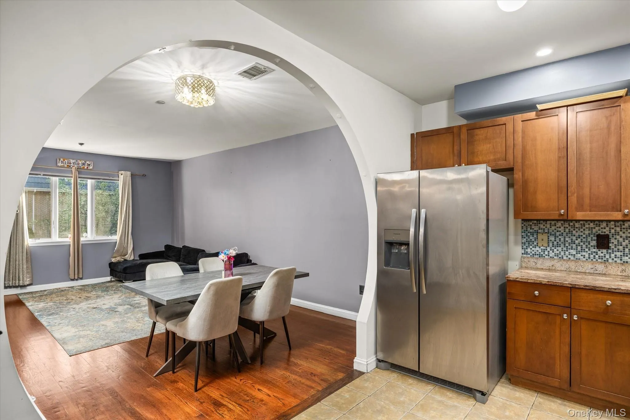 Kitchen and dining area featuring an arched doorway, wood-finish flooring, stainless steel refrigerator, wood cabinetry, and a mosaic tile backsplash Kitchen and dining area featuring an arched doorway, wood-finish flooring, stainless steel refrigerator, wood cabinetry, and a mosaic tile backsplash