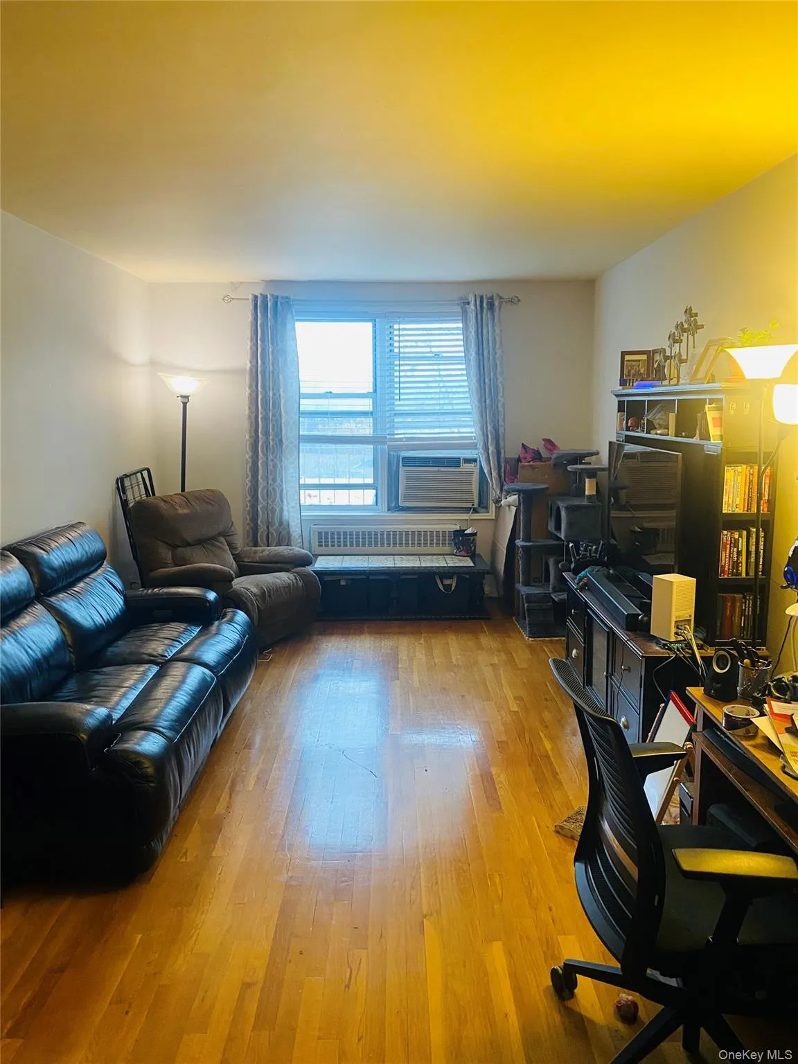 Hardwood flooring throughout a bright room featuring a large window with horizontal blinds and a wall-mounted air conditioning unit Hardwood flooring throughout a bright room featuring a large window with horizontal blinds and a wall-mounted air conditioning unit