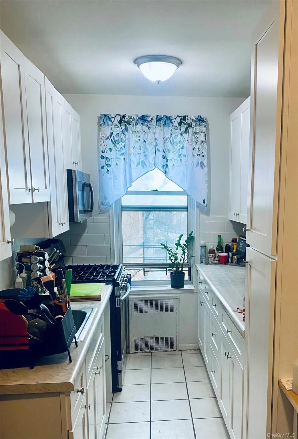Galley kitchen featuring white cabinetry, light-toned countertops, a gas range, stainless steel microwave, and tiled flooring Galley kitchen featuring white cabinetry, light-toned countertops, a gas range, stainless steel microwave, and tiled flooring