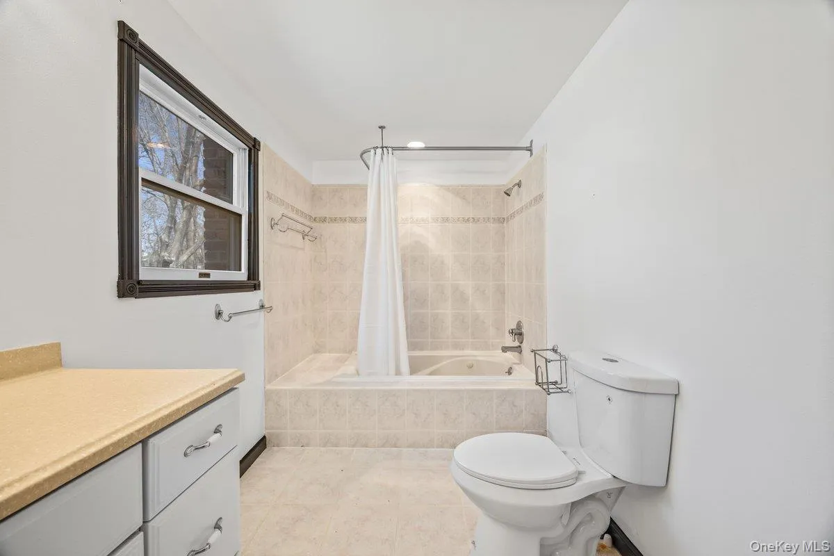 Bathroom featuring a window with dark trim, a tiled bath and shower combination, a toilet, and a vanity with a light-colored countertop Bathroom featuring a window with dark trim, a tiled bath and shower combination, a toilet, and a vanity with a light-colored countertop