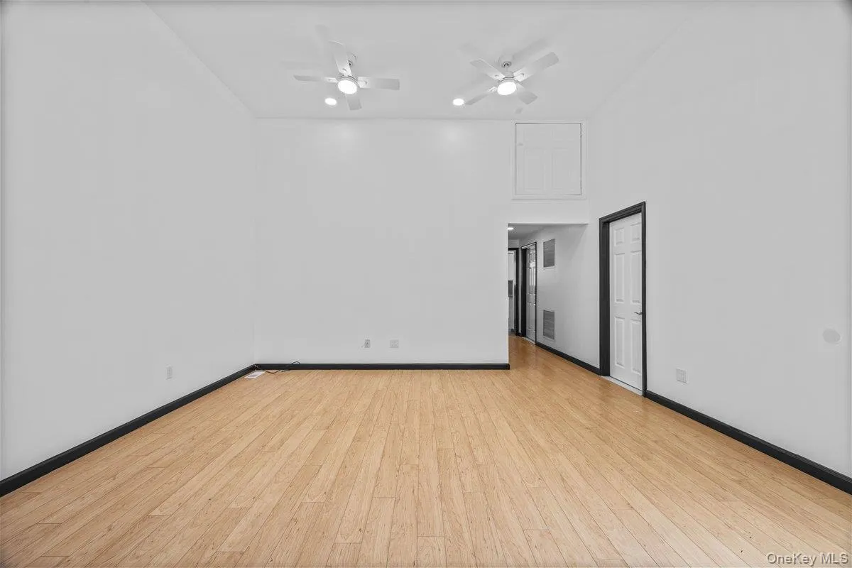 Spacious room featuring light-toned hardwood flooring, white walls, and two ceiling fans with integrated lighting Spacious room featuring light-toned hardwood flooring, white walls, and two ceiling fans with integrated lighting