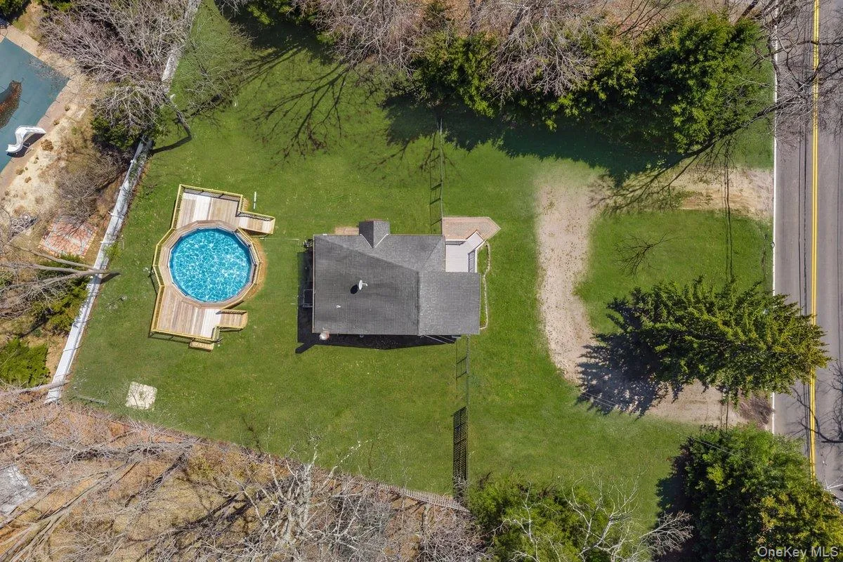 Aerial view of the property featuring a circular pool with an expansive wooden deck, a well-maintained lawn, and mature trees surrounding the perimeter Aerial view of the property featuring a circular pool with an expansive wooden deck, a well-maintained lawn, and mature trees surrounding the perimeter