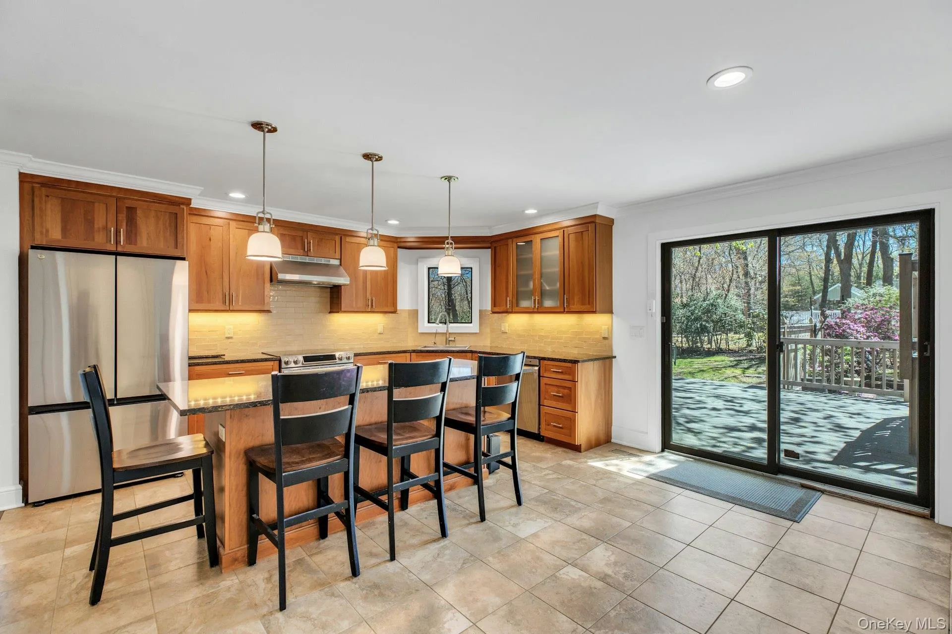 Kitchen featuring wood cabinetry, stainless steel appliances, a center island with pendant lighting, tile flooring, and sliding glass doors opening to an outdoor patio Kitchen featuring wood cabinetry, stainless steel appliances, a center island with pendant lighting, tile flooring, and sliding glass doors opening to an outdoor patio