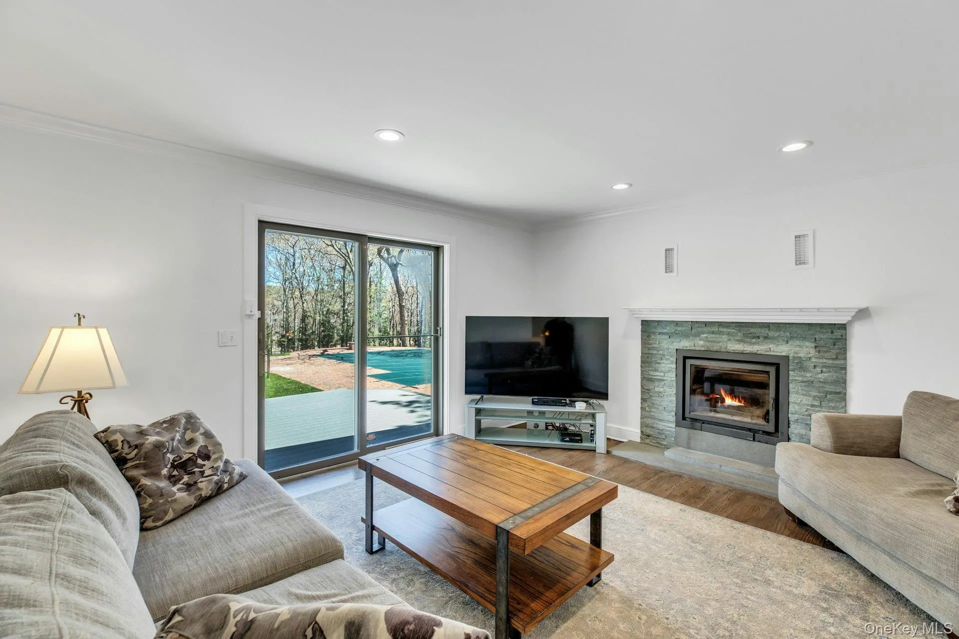 Living area featuring a stone-clad fireplace with a white mantel, recessed lighting, wood-finish flooring, a glass sliding door, and crown molding Living area featuring a stone-clad fireplace with a white mantel, recessed lighting, wood-finish flooring, a glass sliding door, and crown molding