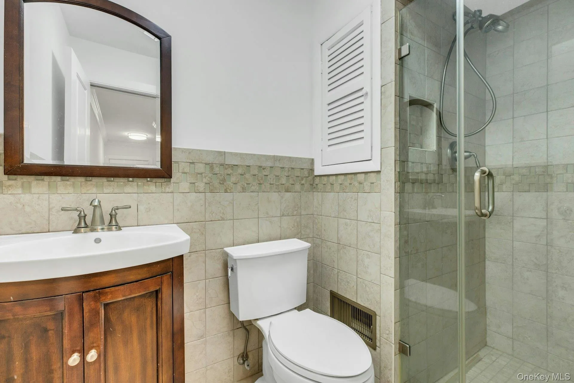 Bathroom featuring a glass-enclosed shower, a wall-mounted vanity with an integrated sink, a dark wood-finish framed mirror, and tiled walls with decorative mosaic accents Bathroom featuring a glass-enclosed shower, a wall-mounted vanity with an integrated sink, a dark wood-finish framed mirror, and tiled walls with decorative mosaic accents