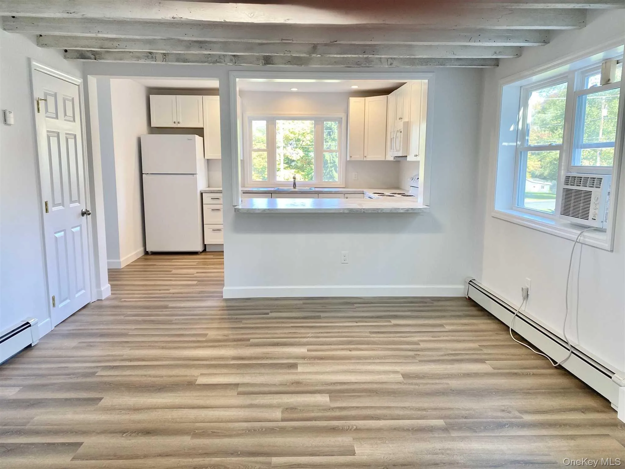 This open-concept living area features light-toned flooring and white walls, complemented by exposed ceiling beams This open-concept living area features light-toned flooring and white walls, complemented by exposed ceiling beams