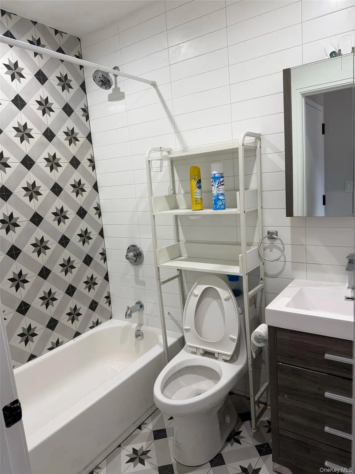 Bathroom featuring patterned floor tiling, a white bathtub with a shower fixture, a toilet, and a vanity with a rectangular sink Bathroom featuring patterned floor tiling, a white bathtub with a shower fixture, a toilet, and a vanity with a rectangular sink