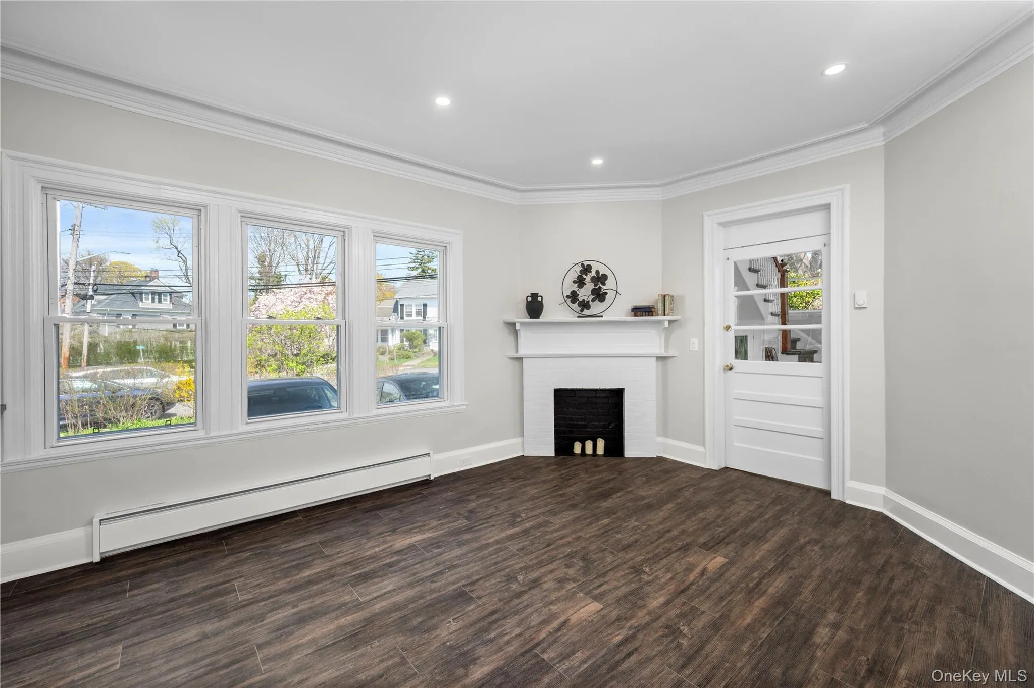 The living area features wood-look flooring, a white mantelpiece above a black decorative fireplace, and three large windows providing extra natural light The living area features wood-look flooring, a white mantelpiece above a black decorative fireplace, and three large windows providing extra natural light
