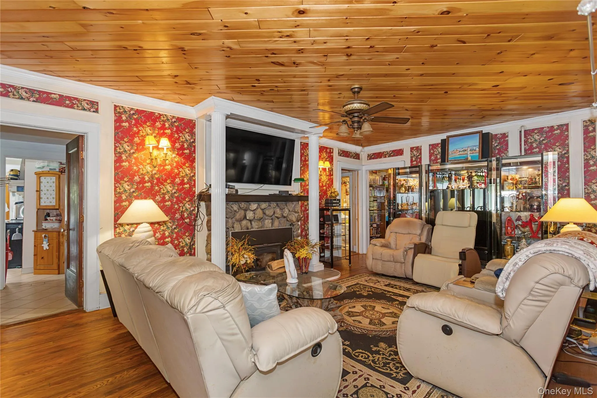 Living room featuring wood ceiling, wood finished floors, ornamental molding, a stone fireplace, and wallpapered walls Living room featuring wood ceiling, wood finished floors, ornamental molding, a stone fireplace, and wallpapered walls