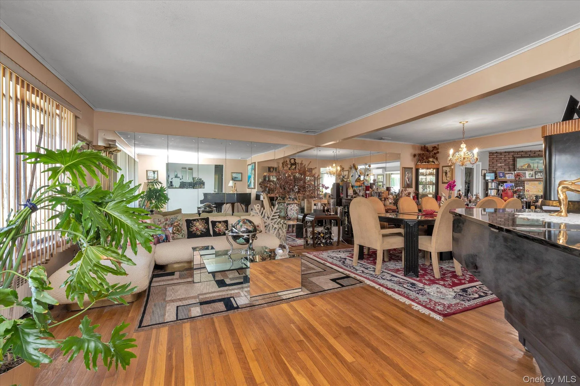 Hardwood flooring extends throughout this expansive living and dining area, featuring mirrored wall accents and a window with vertical blinds Hardwood flooring extends throughout this expansive living and dining area, featuring mirrored wall accents and a window with vertical blinds