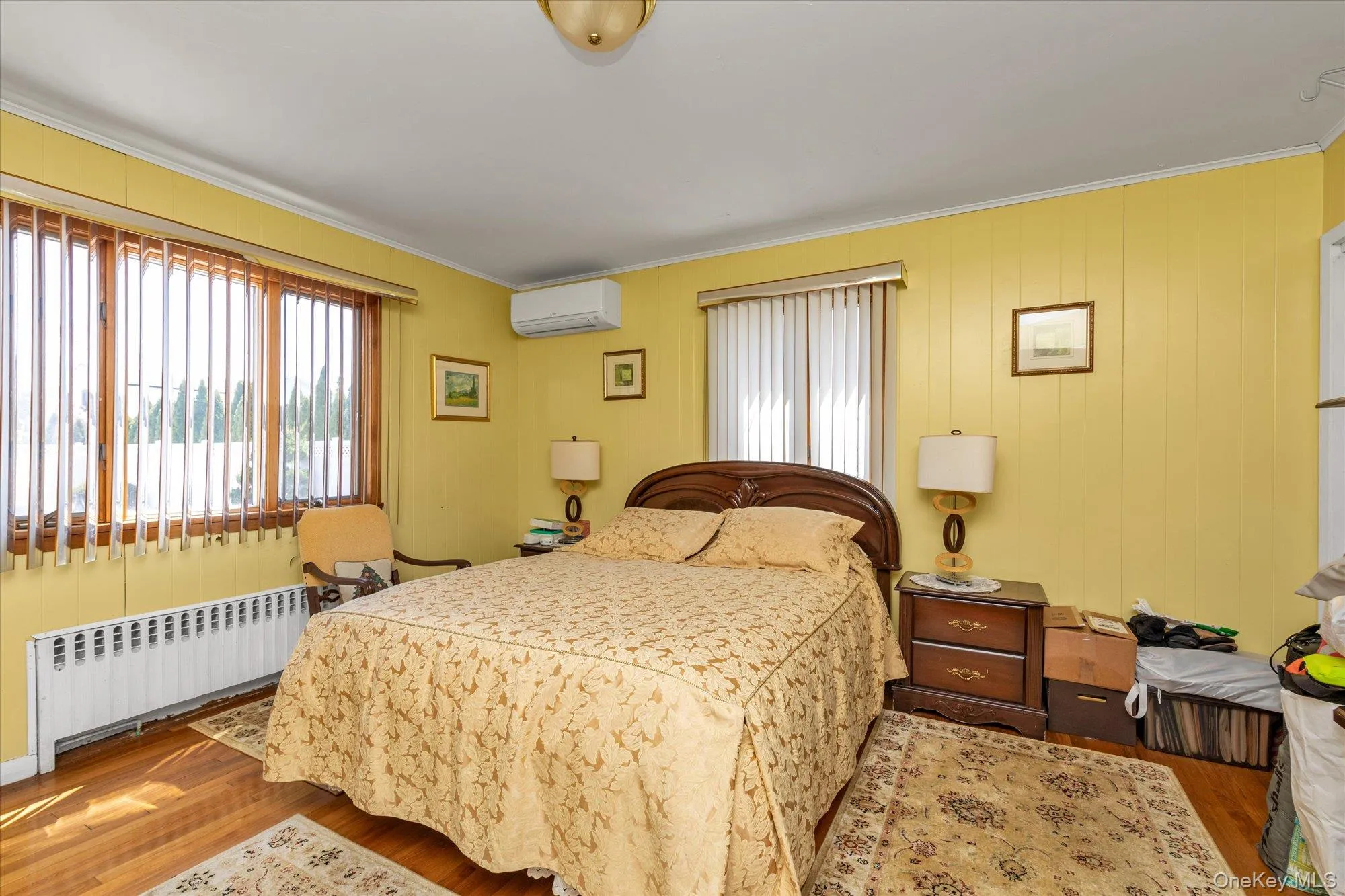 Interior room featuring wood-finish flooring, a wall-mounted AC unit, and a prominent window with vertical blinds-also includes ensuite full bath Interior room featuring wood-finish flooring, a wall-mounted AC unit, and a prominent window with vertical blinds-also includes ensuite full bath