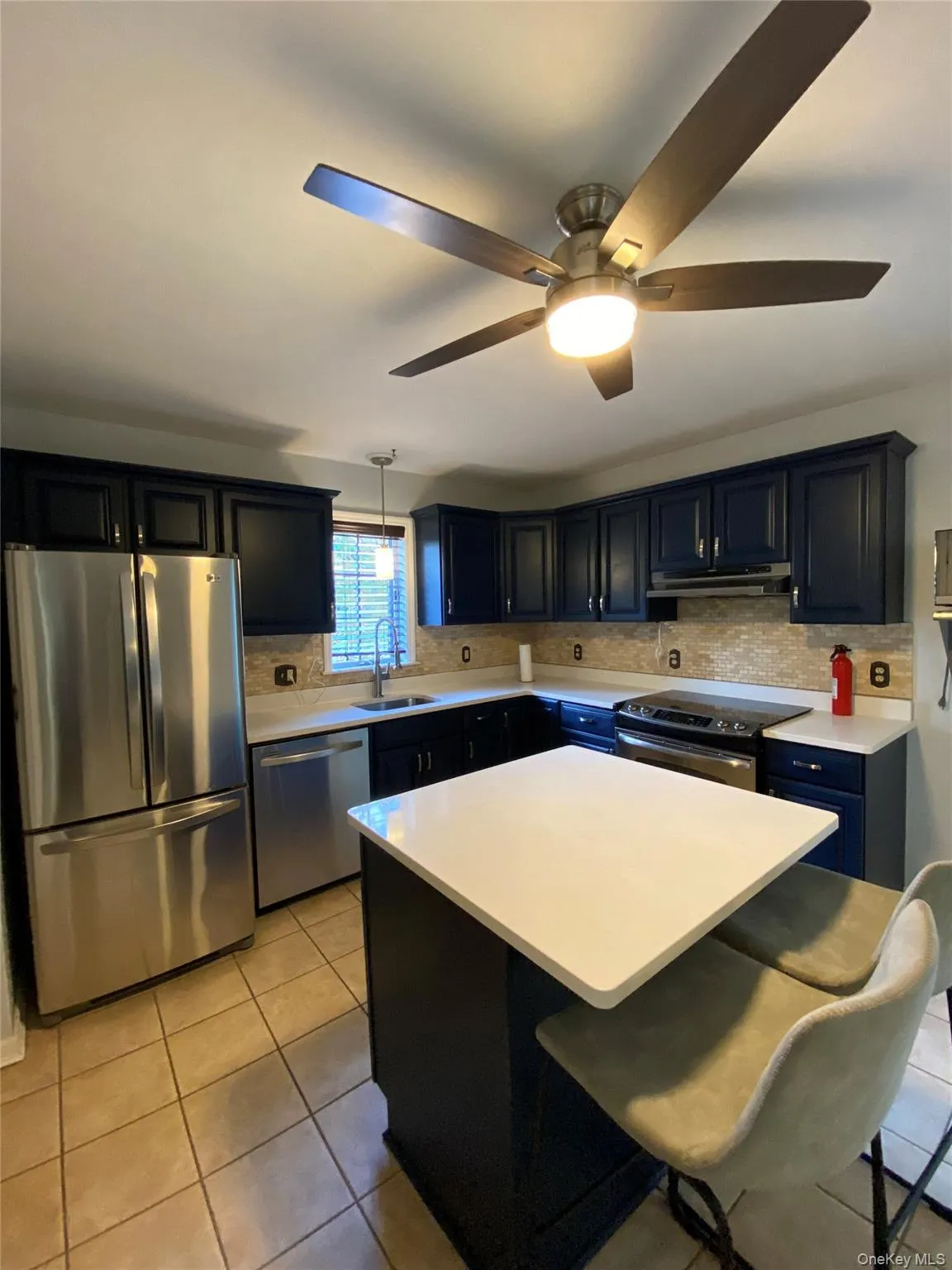 The kitchen features dark cabinetry, stainless steel appliances, a white island countertop, and tiled flooring The kitchen features dark cabinetry, stainless steel appliances, a white island countertop, and tiled flooring
