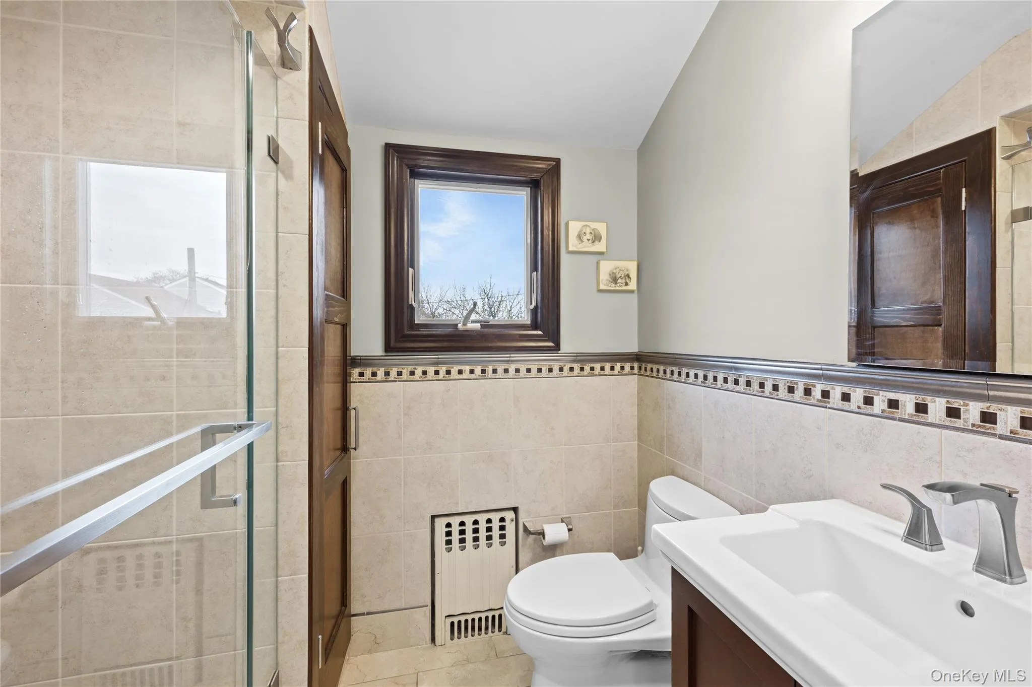 Bathroom featuring a glass-enclosed shower, a vanity with a white sink, and tiled walls with a decorative border Bathroom featuring a glass-enclosed shower, a vanity with a white sink, and tiled walls with a decorative border