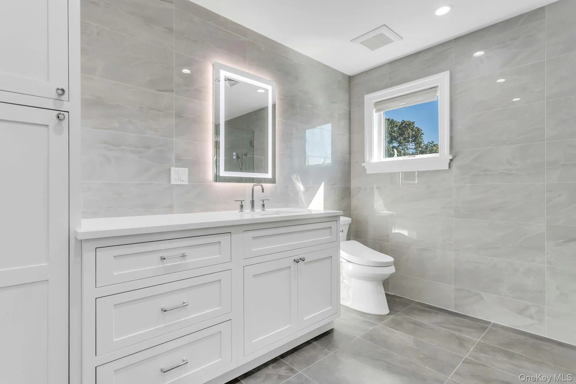 Bathroom featuring a vanity with white cabinetry and a white countertop, an illuminated mirror, and light grey tiled walls and floor Bathroom featuring a vanity with white cabinetry and a white countertop, an illuminated mirror, and light grey tiled walls and floor