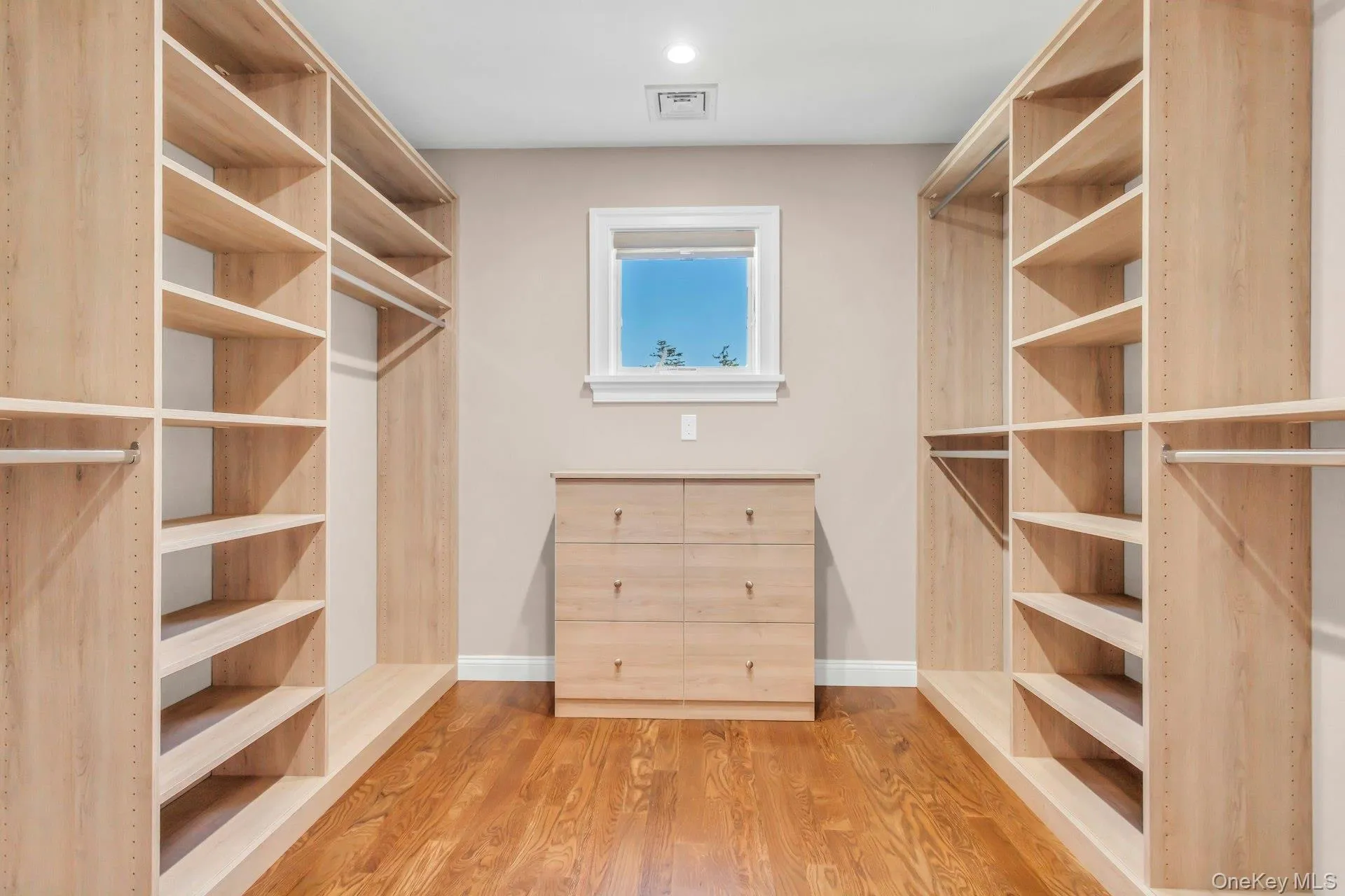 Walk-in closet with extensive built-in shelving, hanging rods, and a central dresser, featuring hardwood floors and a window providing natural light Walk-in closet with extensive built-in shelving, hanging rods, and a central dresser, featuring hardwood floors and a window providing natural light