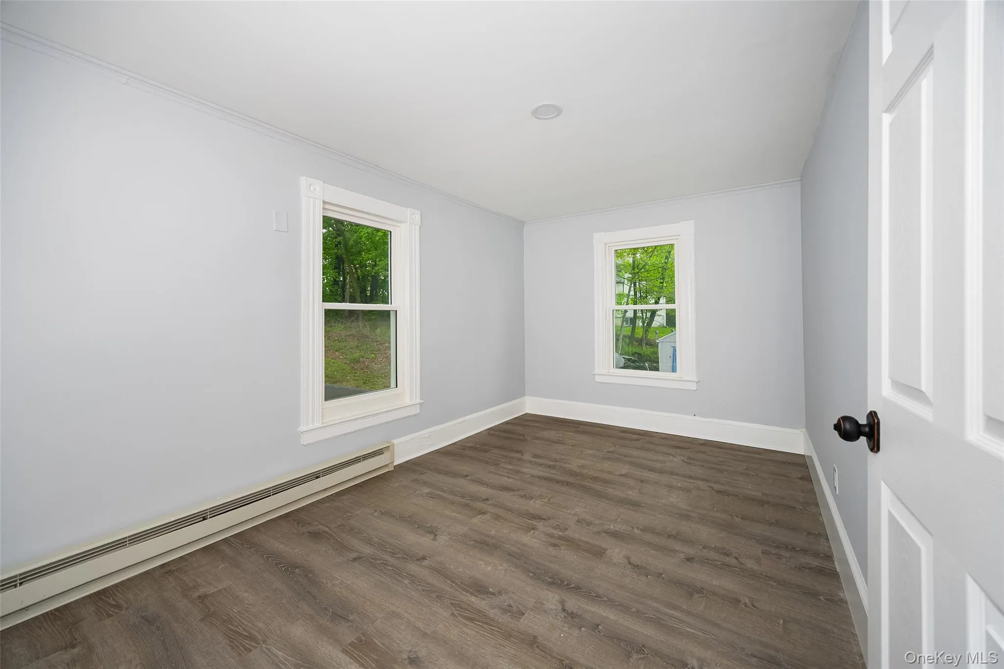 Interior room featuring wood-finish flooring, two window units with white trim, light gray wall paint, and white baseboards Interior room featuring wood-finish flooring, two window units with white trim, light gray wall paint, and white baseboards