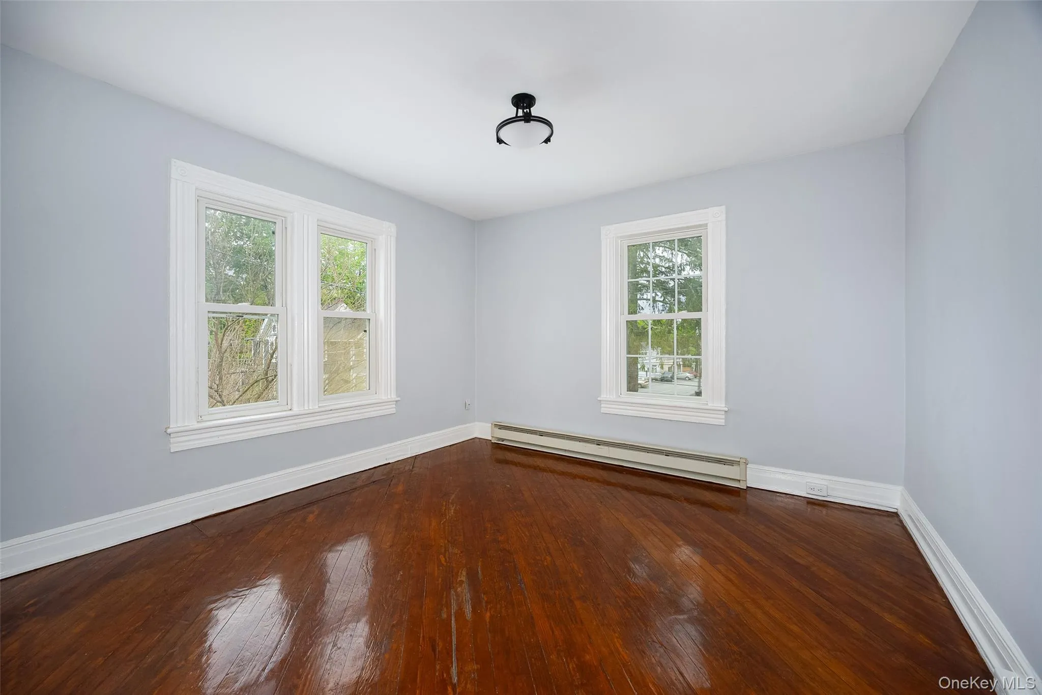 Room featuring rich wood flooring, light gray walls, multiple windows with white trim, white baseboards, and a semi-flush mount ceiling light fixture Room featuring rich wood flooring, light gray walls, multiple windows with white trim, white baseboards, and a semi-flush mount ceiling light fixture