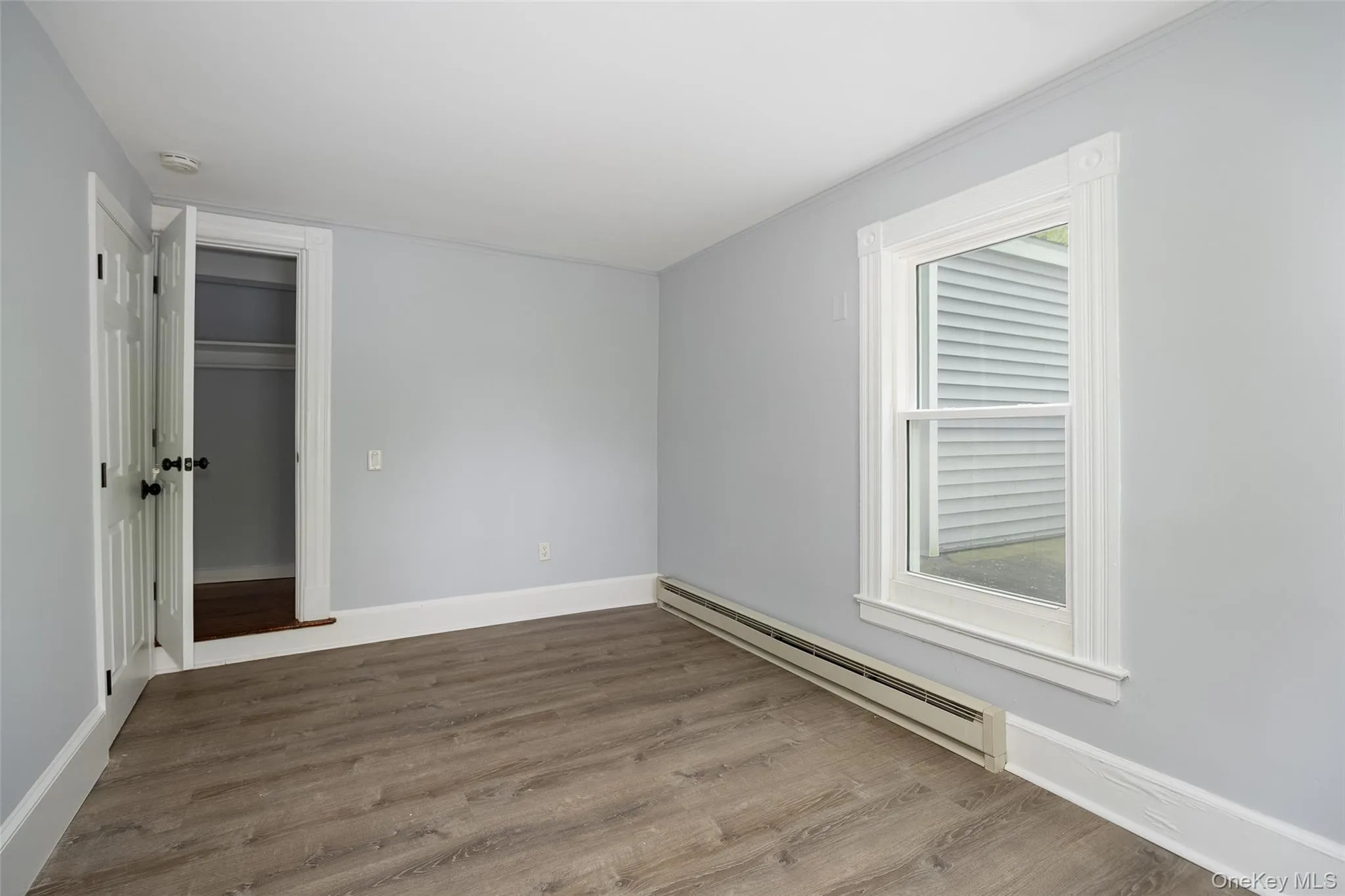 Room featuring wood-finish flooring, a window with decorative trim, a baseboard heater, and a closet with bifold doors Room featuring wood-finish flooring, a window with decorative trim, a baseboard heater, and a closet with bifold doors