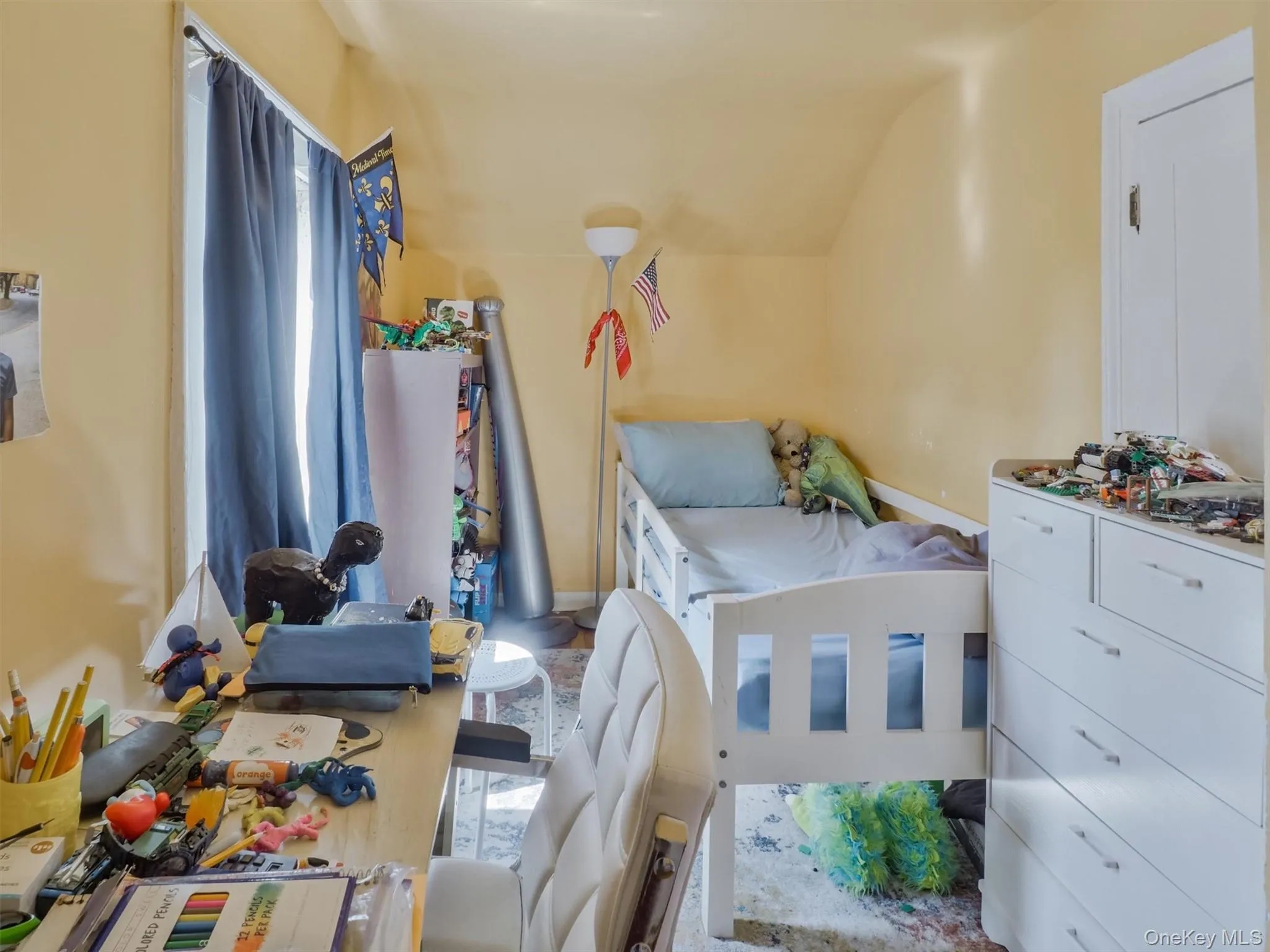 This room features a window with blue curtains, a bed with a white frame, and a white dresser with drawers This room features a window with blue curtains, a bed with a white frame, and a white dresser with drawers