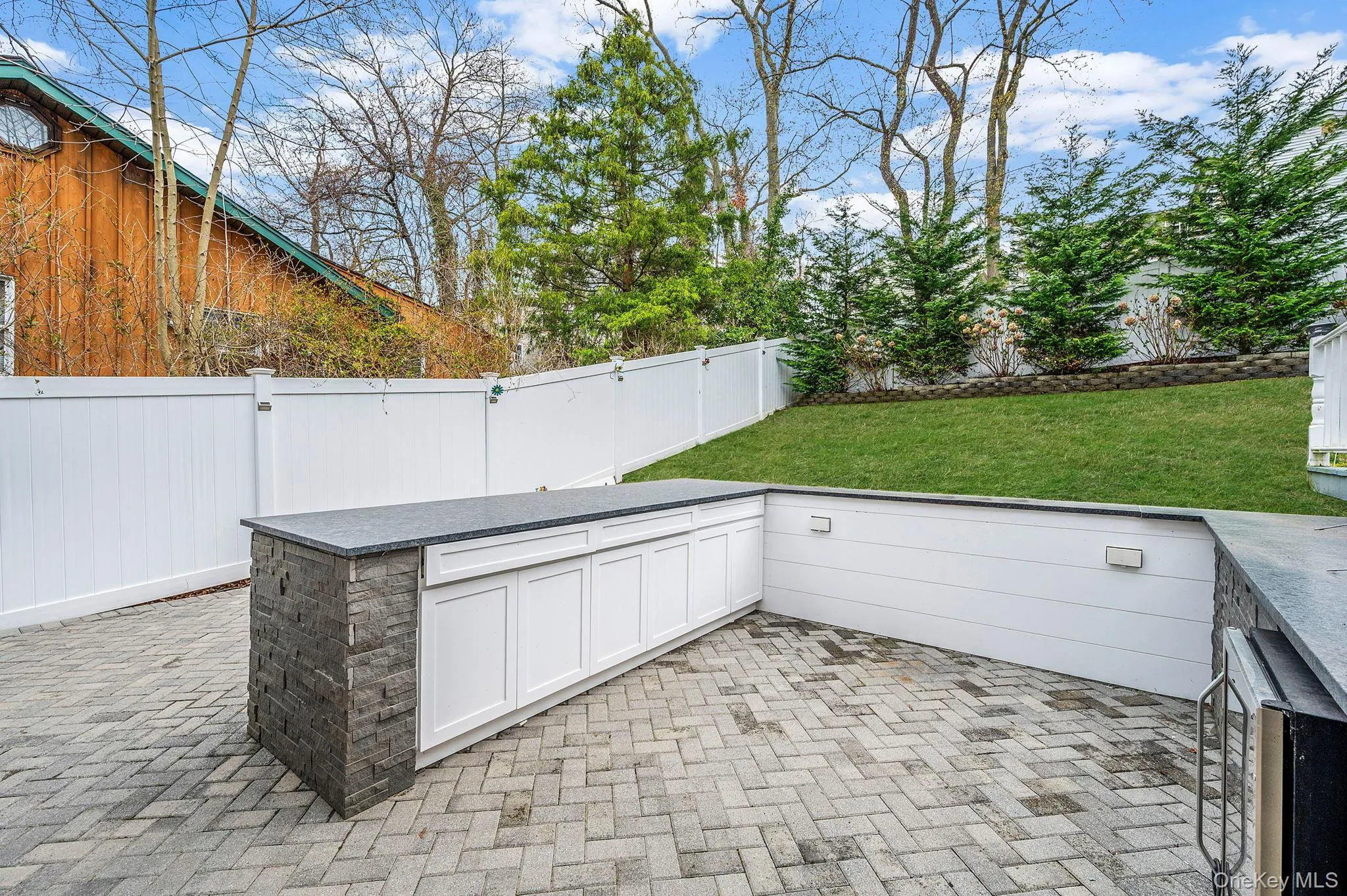 The property features an outdoor entertaining area with a paved patio, a built-in outdoor kitchen with a stone facade and white cabinetry, and a landscaped yard with a lawn and evergreen trees The property features an outdoor entertaining area with a paved patio, a built-in outdoor kitchen with a stone facade and white cabinetry, and a landscaped yard with a lawn and evergreen trees