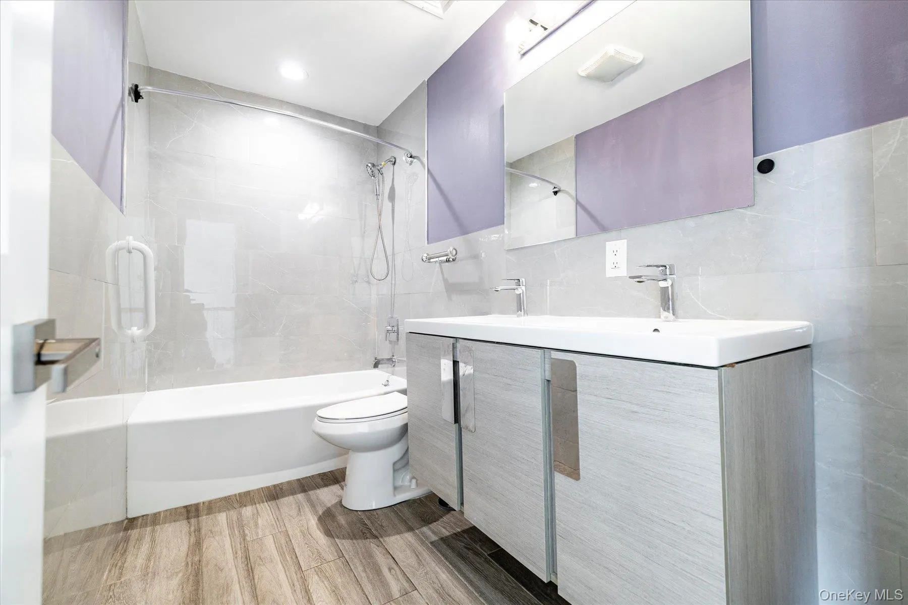 Bathroom featuring a dual-sink vanity, wood-look flooring, large wall mirror, and a bathtub with a tiled surround Bathroom featuring a dual-sink vanity, wood-look flooring, large wall mirror, and a bathtub with a tiled surround