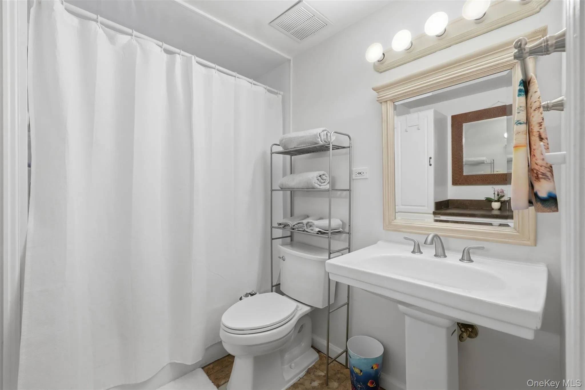 Bathroom featuring a white pedestal sink with brushed nickel fixtures, a framed mirror with vanity lighting, and a white shower curtain concealing a tub/shower combination Bathroom featuring a white pedestal sink with brushed nickel fixtures, a framed mirror with vanity lighting, and a white shower curtain concealing a tub/shower combination