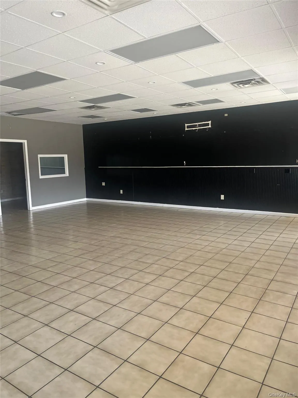 Spacious room featuring light-colored square floor tiles, a drop ceiling with recessed lighting, and a dark accent wall Spacious room featuring light-colored square floor tiles, a drop ceiling with recessed lighting, and a dark accent wall