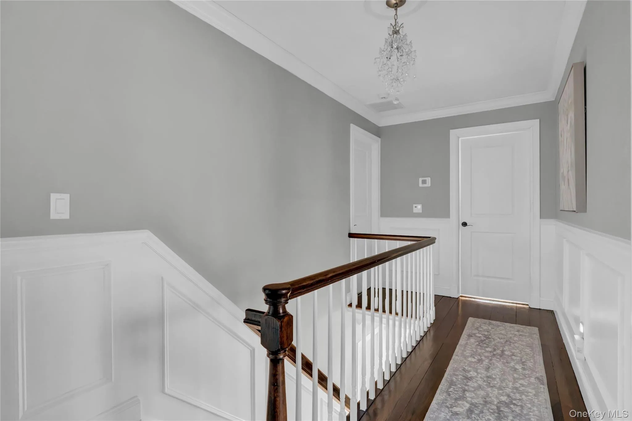 Upper-level landing featuring wainscoting, crown molding, dark wood-finish flooring, and a crystal chandelier Upper-level landing featuring wainscoting, crown molding, dark wood-finish flooring, and a crystal chandelier