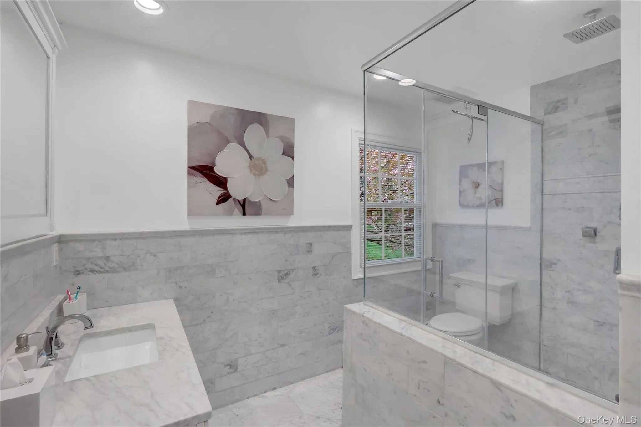 Bathroom featuring extensive white stone tile, a glass-enclosed shower, a large vanity with an integrated sink, a window with natural light, and recessed lighting Bathroom featuring extensive white stone tile, a glass-enclosed shower, a large vanity with an integrated sink, a window with natural light, and recessed lighting