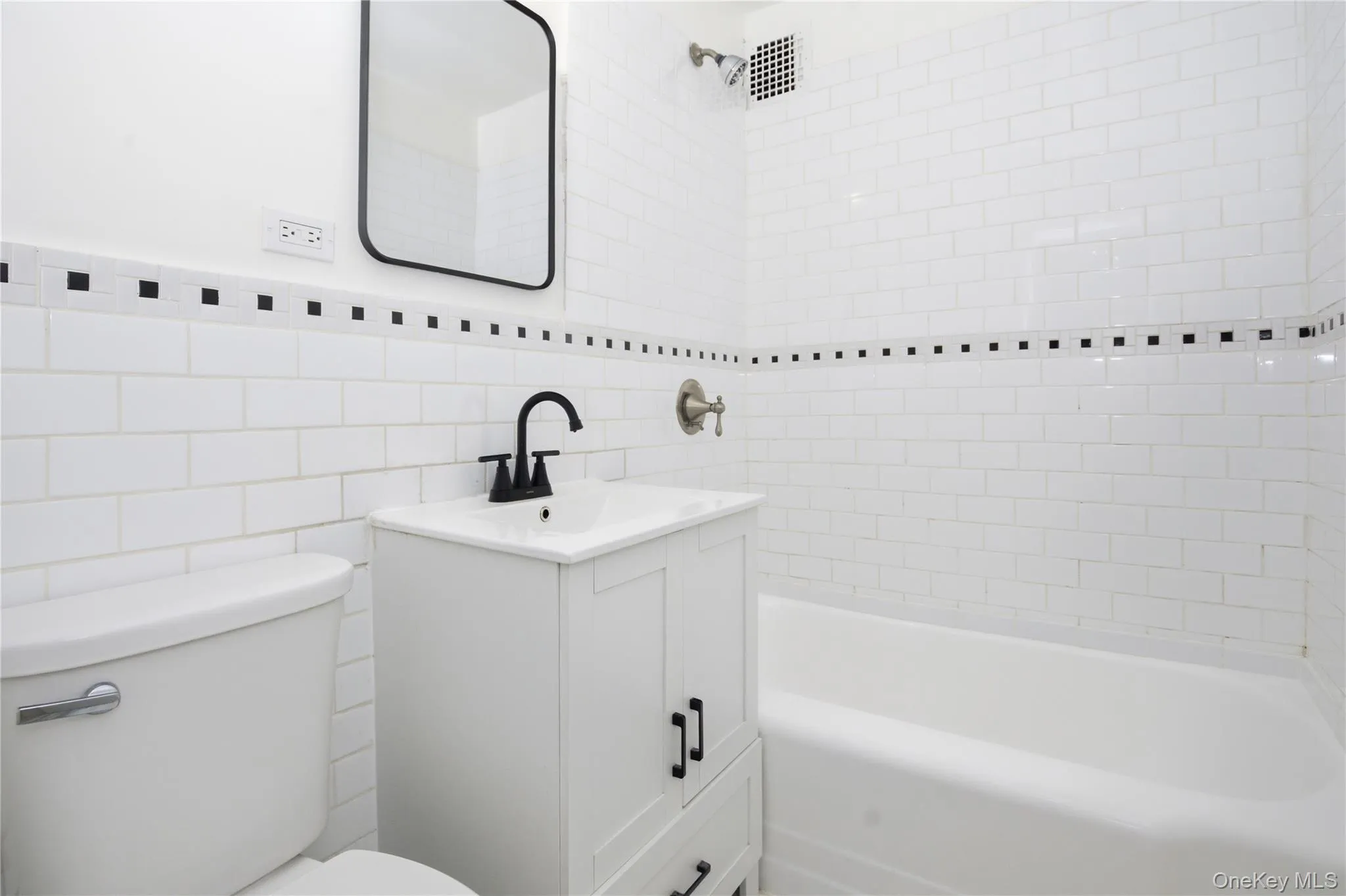 Bathroom featuring white subway tile walls with a decorative black tile accent, a white vanity with a dark faucet, a white toilet, and a white bathtub with a shower head Bathroom featuring white subway tile walls with a decorative black tile accent, a white vanity with a dark faucet, a white toilet, and a white bathtub with a shower head