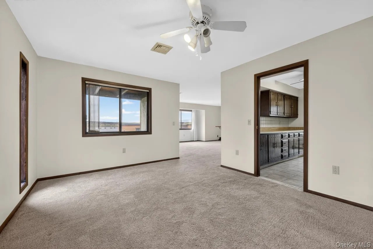 Carpeted living area featuring a ceiling fan with light fixture, large window with dark trim, and a doorway opening to a kitchen with dark wood cabinetry Carpeted living area featuring a ceiling fan with light fixture, large window with dark trim, and a doorway opening to a kitchen with dark wood cabinetry