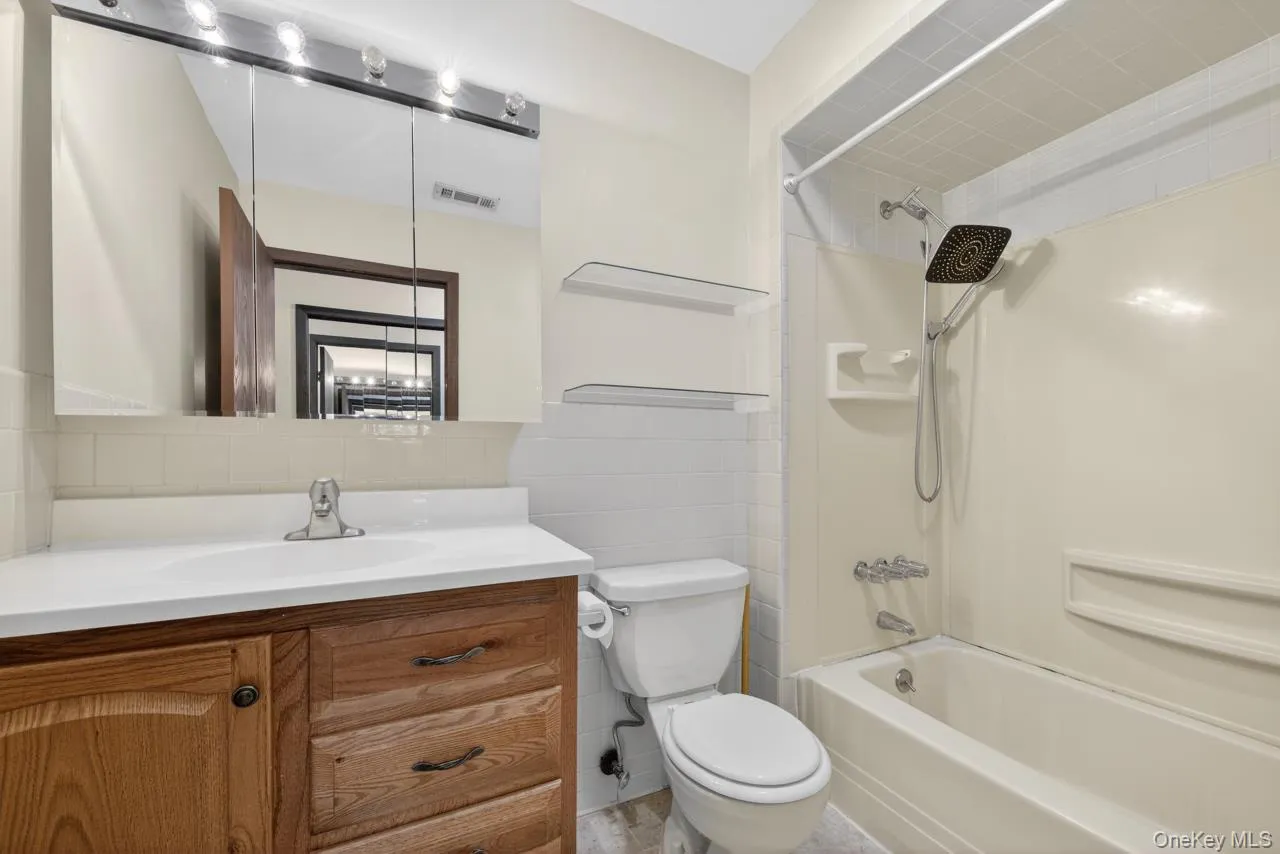 Bathroom featuring a wood-finish vanity with a white countertop and integrated sink, a mirrored medicine cabinet with overhead lighting, a toilet, and a white tub/shower combination with a flexible showerhead Bathroom featuring a wood-finish vanity with a white countertop and integrated sink, a mirrored medicine cabinet with overhead lighting, a toilet, and a white tub/shower combination with a flexible showerhead