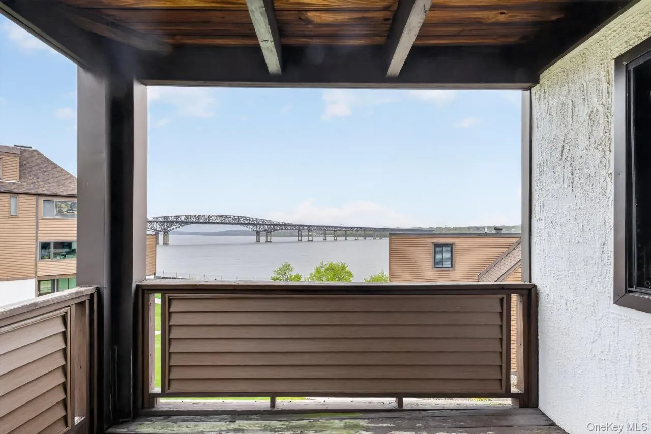 Balcony featuring a wood-panel ceiling and dark railing, overlooking a body of water with a bridge in the distance Balcony featuring a wood-panel ceiling and dark railing, overlooking a body of water with a bridge in the distance