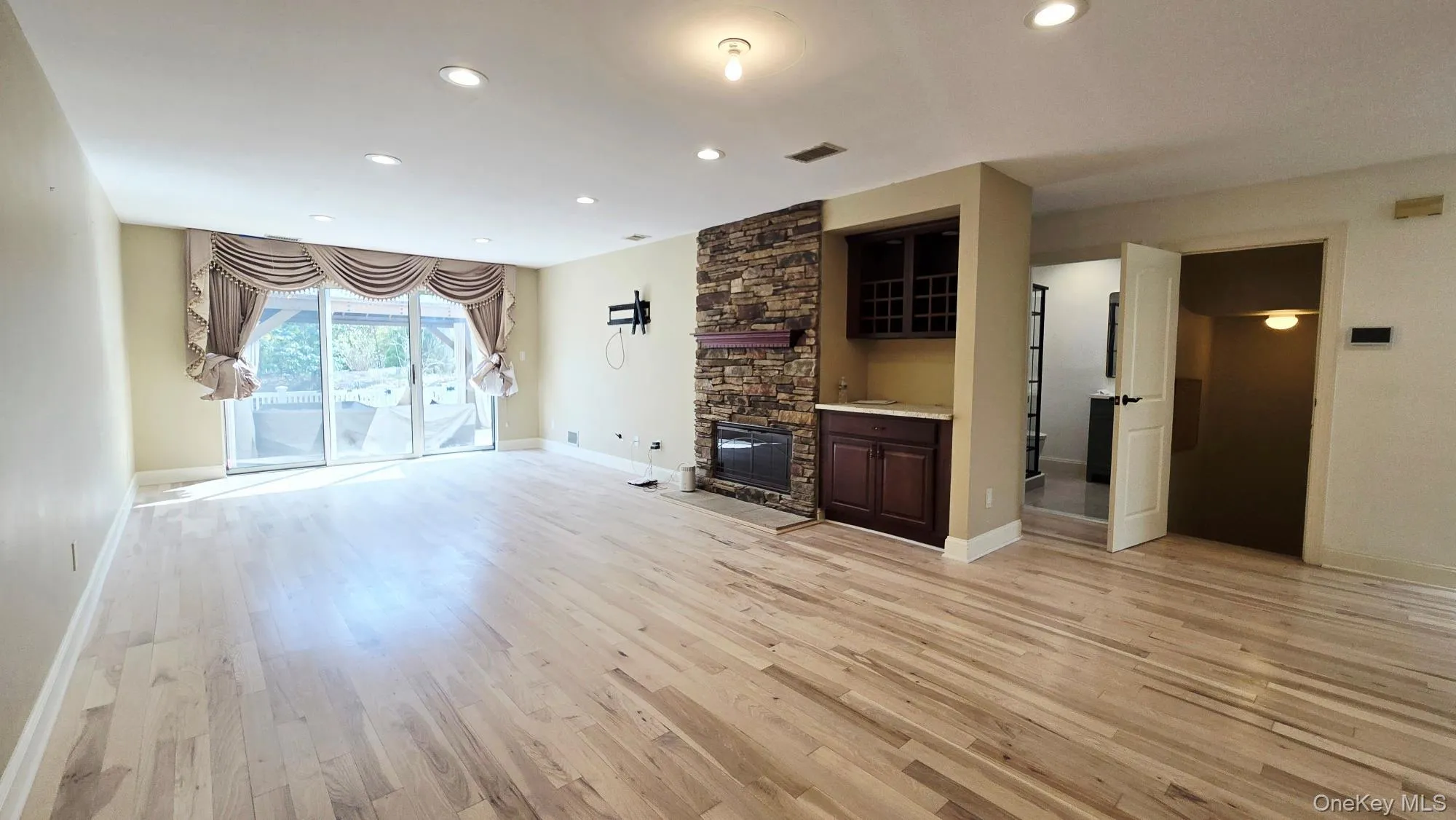 Spacious living area featuring light-toned hardwood flooring, recessed lighting, and sliding glass doors with draped valances Spacious living area featuring light-toned hardwood flooring, recessed lighting, and sliding glass doors with draped valances