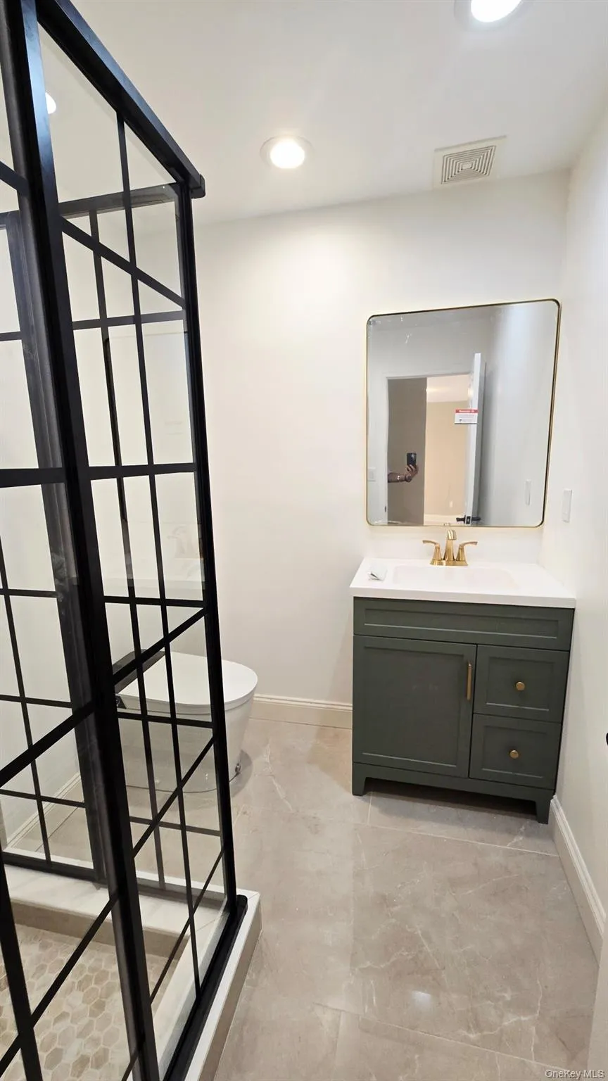 The full bathroom on the first floor features a walk-in shower with a black grid-patterned glass enclosure, a vanity with a dark green cabinet and gold hardware, and a square-edged mirror The full bathroom on the first floor features a walk-in shower with a black grid-patterned glass enclosure, a vanity with a dark green cabinet and gold hardware, and a square-edged mirror