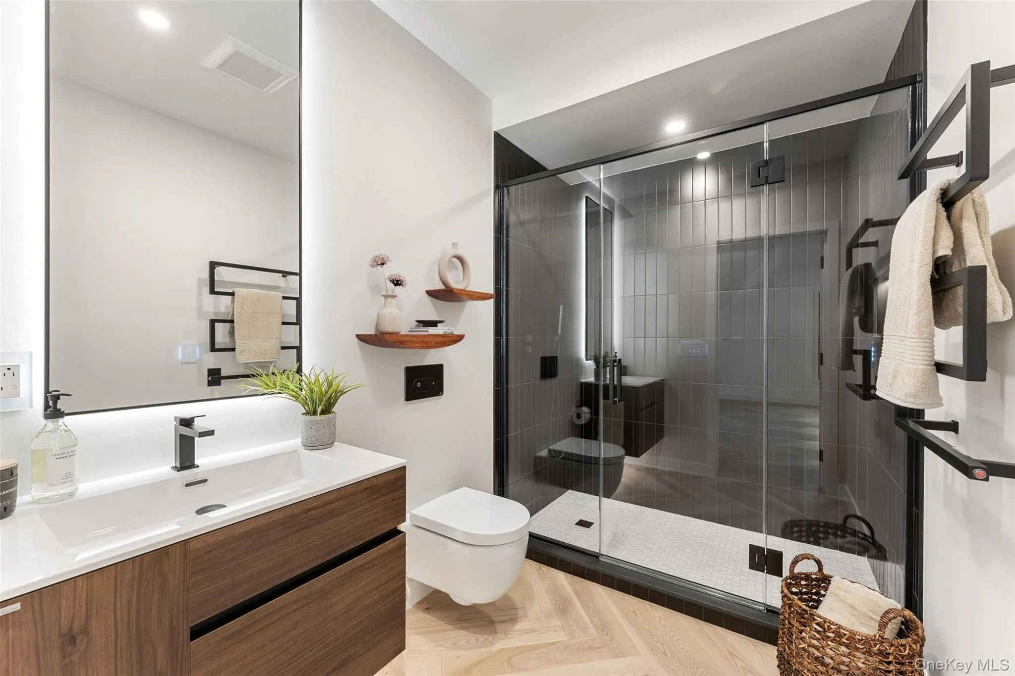 Modern ensuite bathroom in 2nd bedroom featuring a wood vanity with a white countertop, a rectangular mirror with backlighting, a glass-enclosed shower with dark tile, and light-colored flooring Modern ensuite bathroom in 2nd bedroom featuring a wood vanity with a white countertop, a rectangular mirror with backlighting, a glass-enclosed shower with dark tile, and light-colored flooring