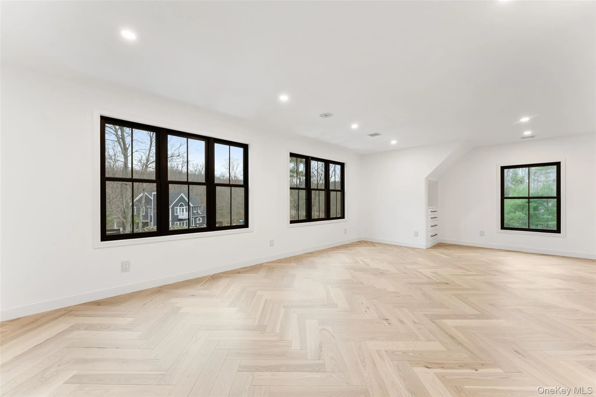 Expansive bedroom featuring light hardwood floors laid in a herringbone pattern, recessed lighting, and multiple windows with dark frames Expansive bedroom featuring light hardwood floors laid in a herringbone pattern, recessed lighting, and multiple windows with dark frames