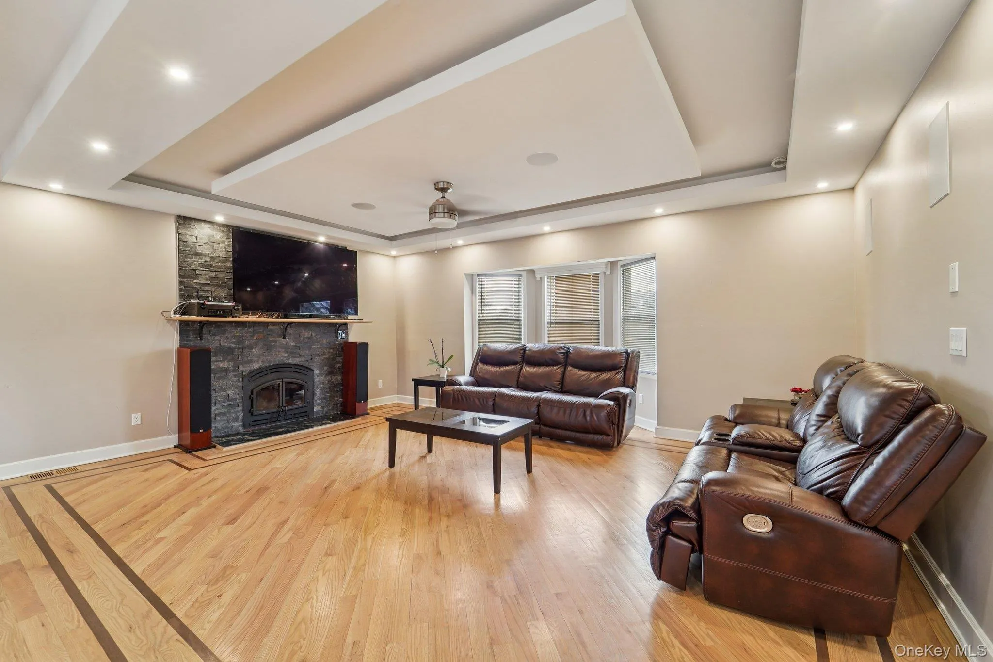 Living area featuring hardwood flooring with decorative inlay, a stone-clad fireplace, and tray ceilings with recessed lighting. Living area featuring hardwood flooring with decorative inlay, a stone-clad fireplace, and tray ceilings with recessed lighting.