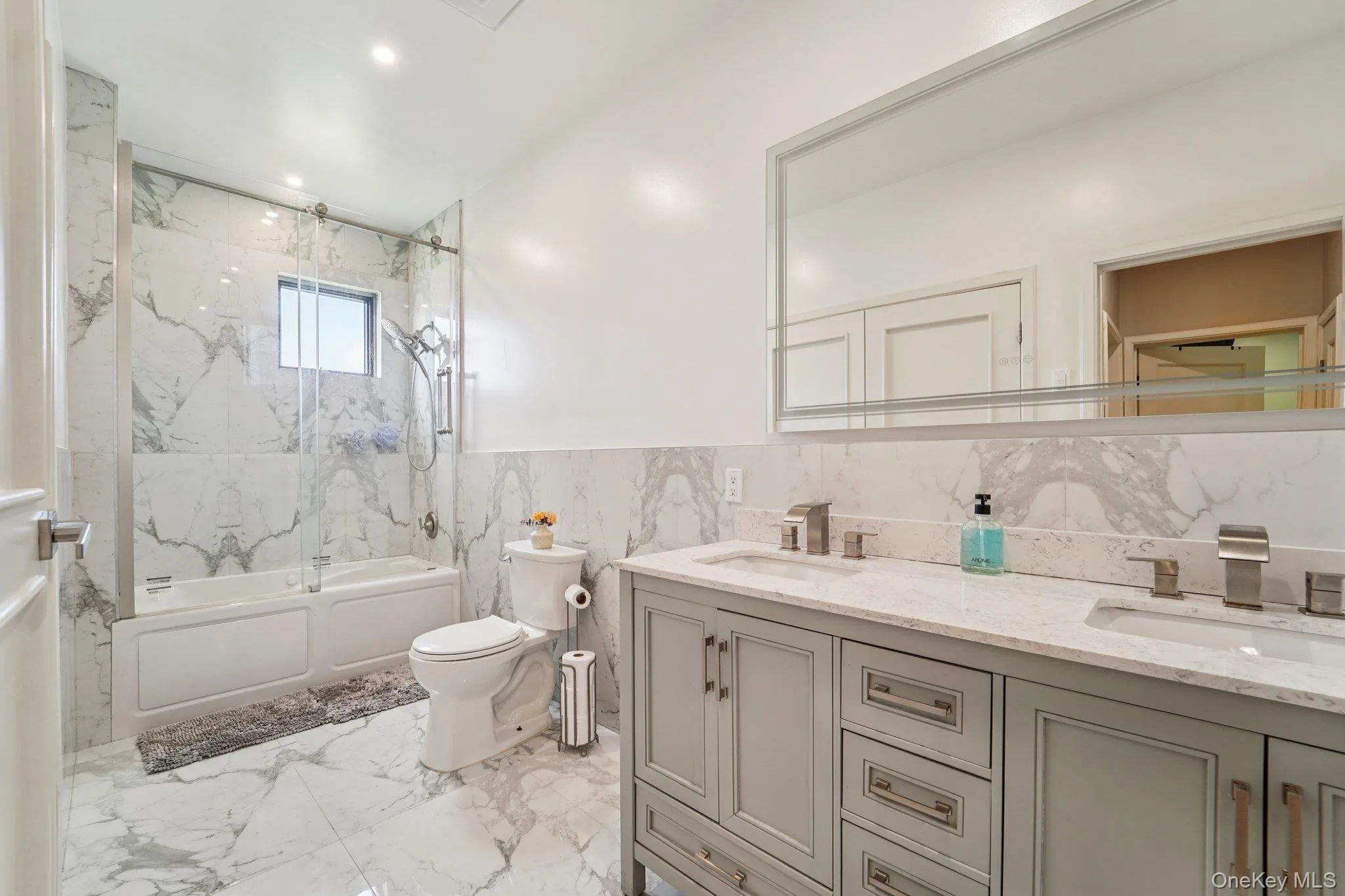 Bathroom 2: This bathroom features a double vanity with light gray cabinetry and brushed nickel hardware, set against a backdrop of white marble-patterned tile flooring and wall accents. Includes laundry area. Bathroom 2: This bathroom features a double vanity with light gray cabinetry and brushed nickel hardware, set against a backdrop of white marble-patterned tile flooring and wall accents. Includes laundry area.