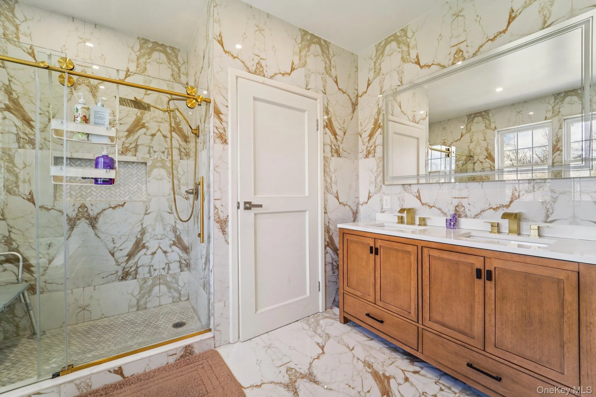This bathroom features elegant marble-patterned tile flooring and walls, a spacious shower with brass-toned fixtures and a glass enclosure, and a double vanity with a wooden cabinet and white countertops. This bathroom features elegant marble-patterned tile flooring and walls, a spacious shower with brass-toned fixtures and a glass enclosure, and a double vanity with a wooden cabinet and white countertops.