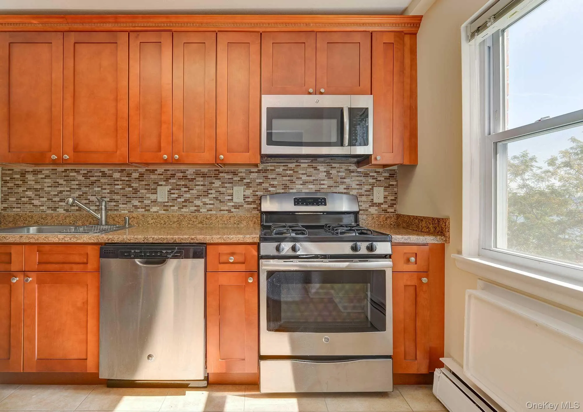 The kitchen features natural wood cabinetry, a mosaic tile backsplash, stainless steel appliances including a gas range, and a window providing natural light The kitchen features natural wood cabinetry, a mosaic tile backsplash, stainless steel appliances including a gas range, and a window providing natural light