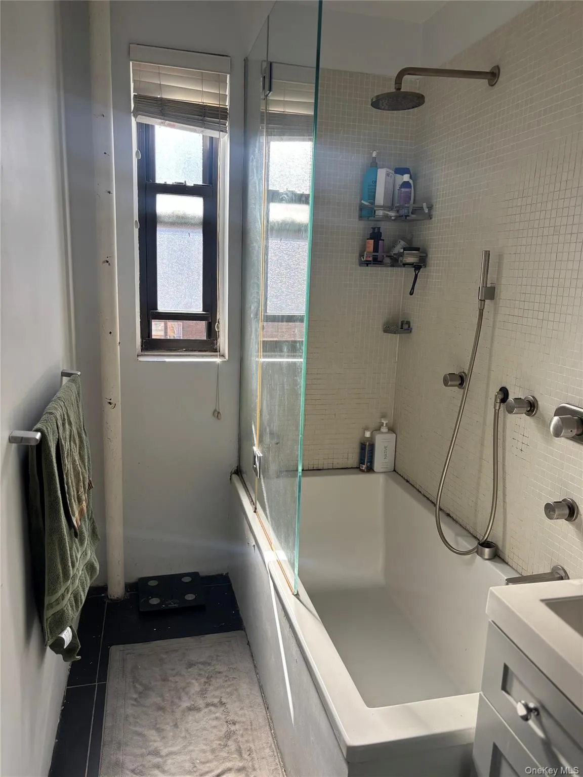 The bathroom features a bathtub with a glass shower screen, a wall-mounted showerhead, and a window with blinds The bathroom features a bathtub with a glass shower screen, a wall-mounted showerhead, and a window with blinds