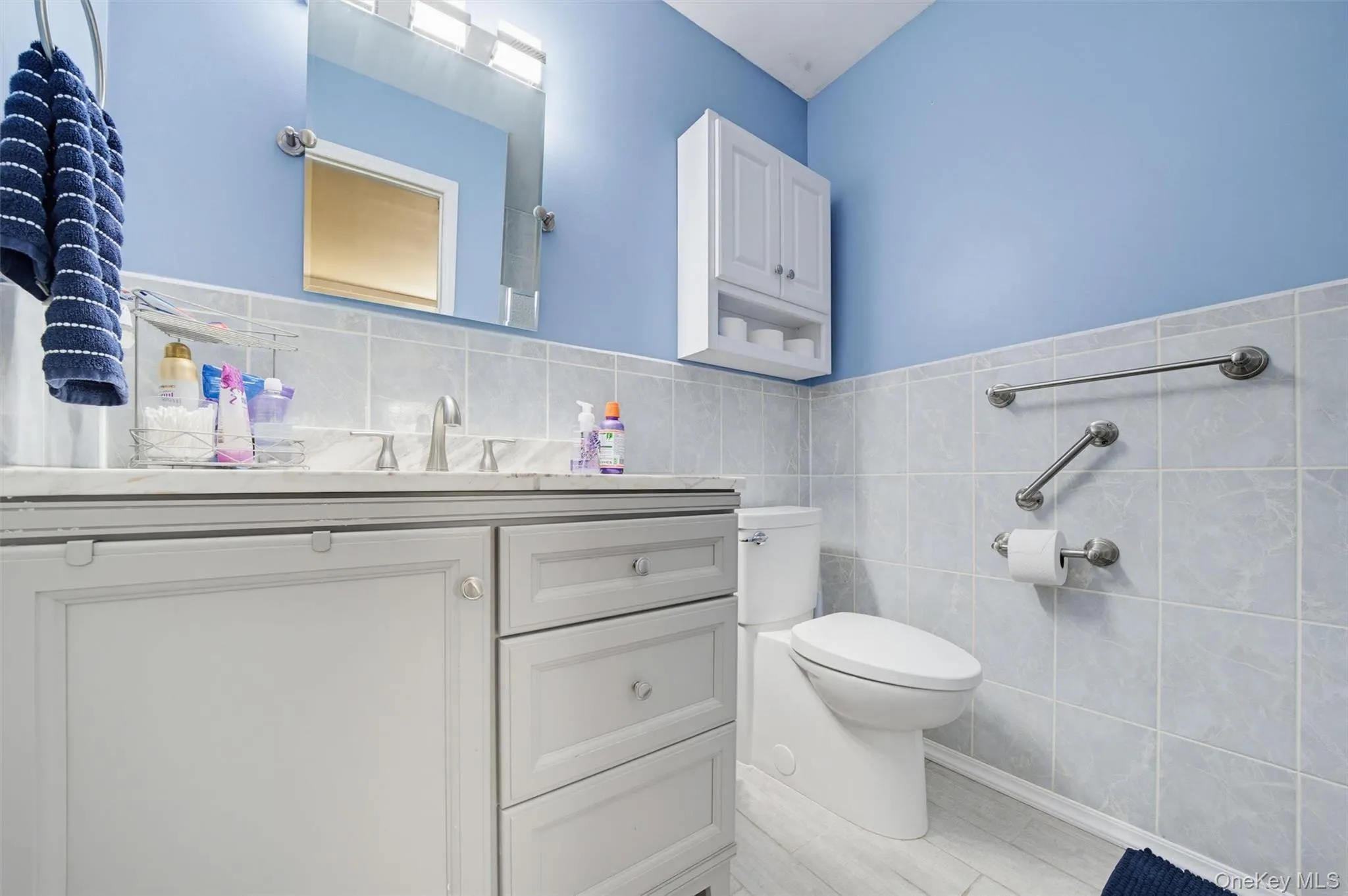 Bathroom featuring a vanity with a white countertop and a mirror above, a toilet, a wall-mounted cabinet, and light blue walls with tiled wainscoting Bathroom featuring a vanity with a white countertop and a mirror above, a toilet, a wall-mounted cabinet, and light blue walls with tiled wainscoting