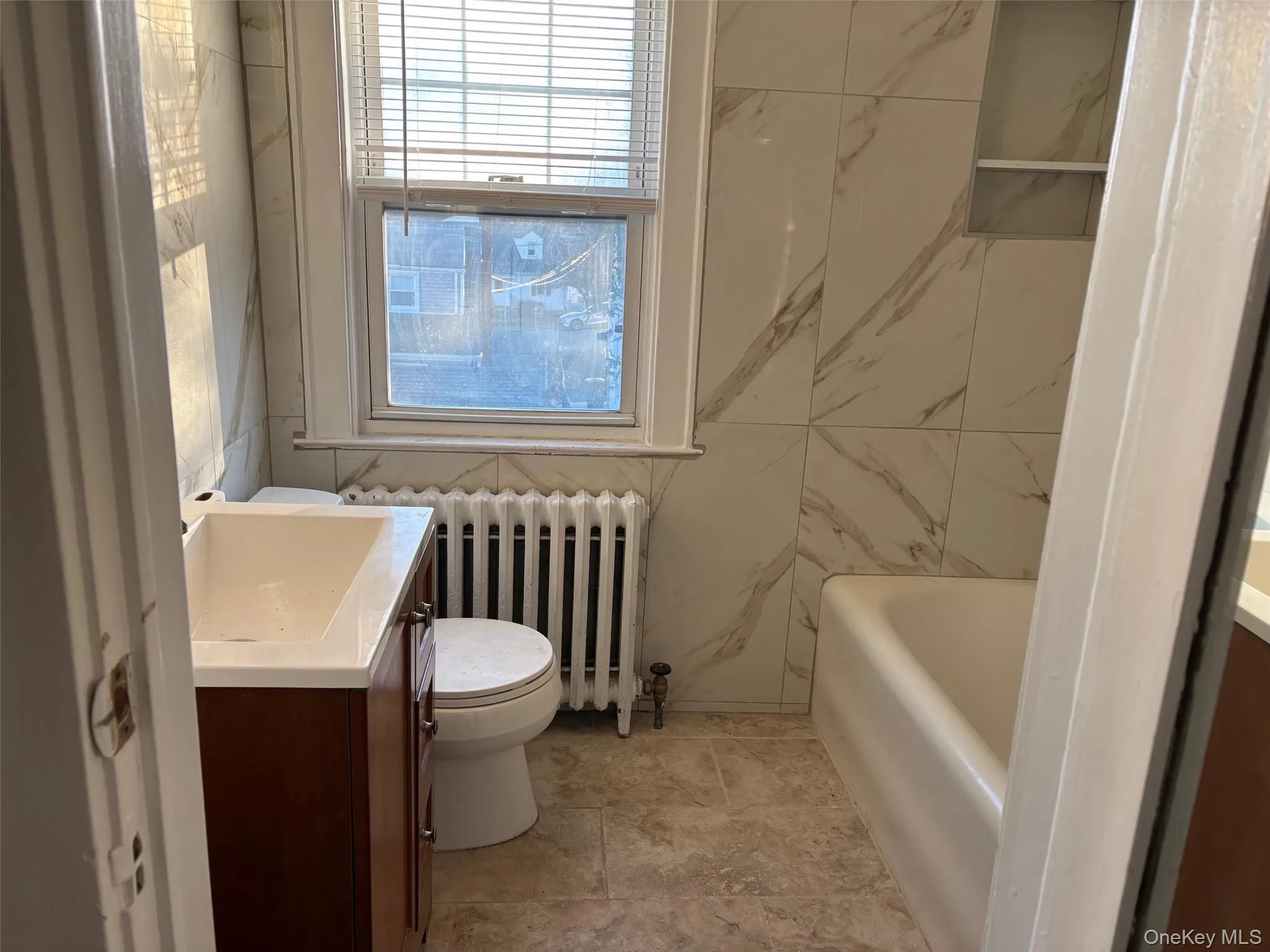 The bathroom features a vanity with an integrated sink, a toilet, a window with blinds, and a bathtub The bathroom features a vanity with an integrated sink, a toilet, a window with blinds, and a bathtub