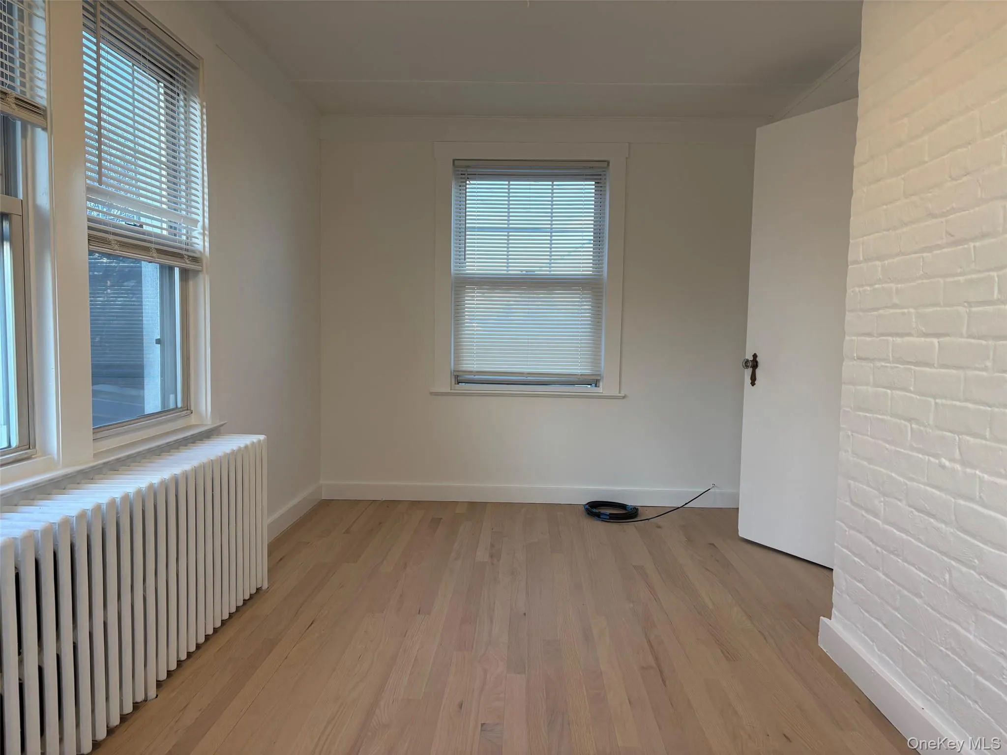 Bright room featuring light-toned hardwood floors, two windows with blinds, a white radiator, and a white brick wall accent Bright room featuring light-toned hardwood floors, two windows with blinds, a white radiator, and a white brick wall accent