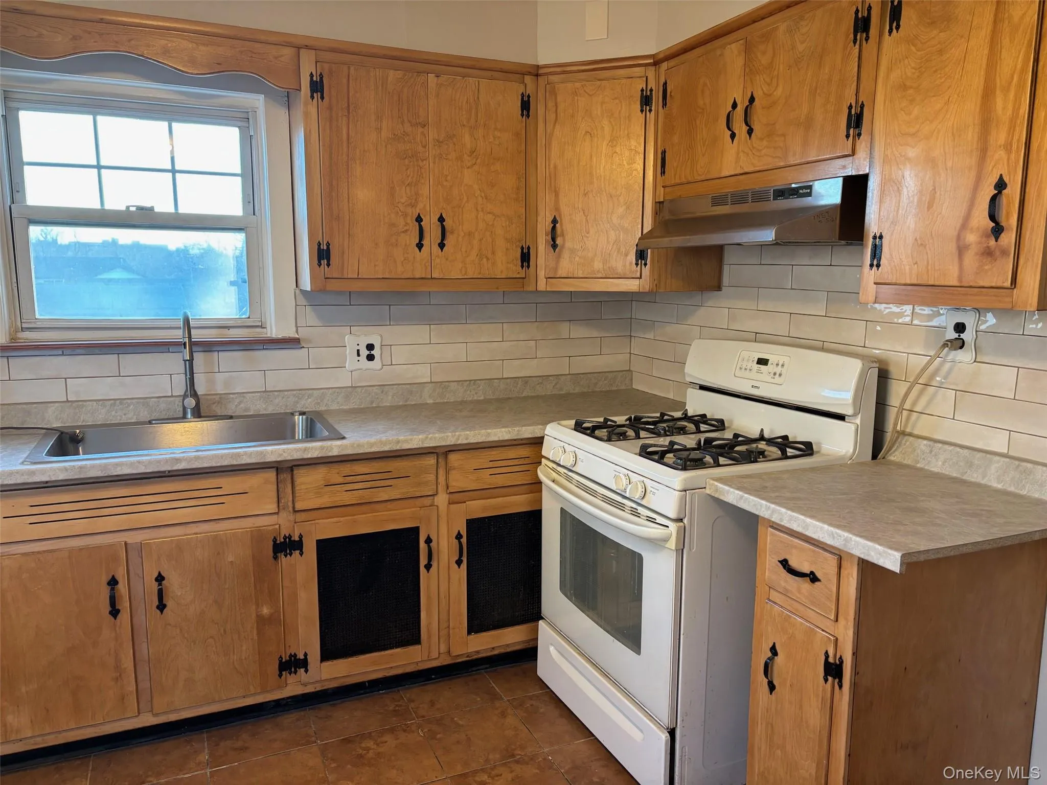 The kitchen features wood cabinetry with black hardware, a stainless steel sink, a white gas range with an oven, and a tiled backsplash The kitchen features wood cabinetry with black hardware, a stainless steel sink, a white gas range with an oven, and a tiled backsplash