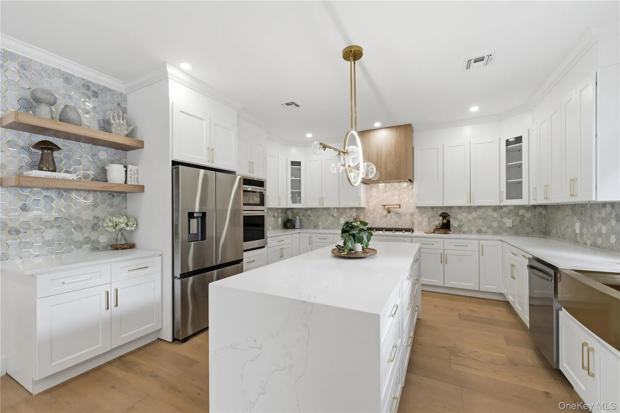 Modern kitchen featuring an expansive central island with a waterfall countertop, white shaker-style cabinetry, stainless steel appliances, a mosaic tile backsplash, and light wood-finish flooring Modern kitchen featuring an expansive central island with a waterfall countertop, white shaker-style cabinetry, stainless steel appliances, a mosaic tile backsplash, and light wood-finish flooring