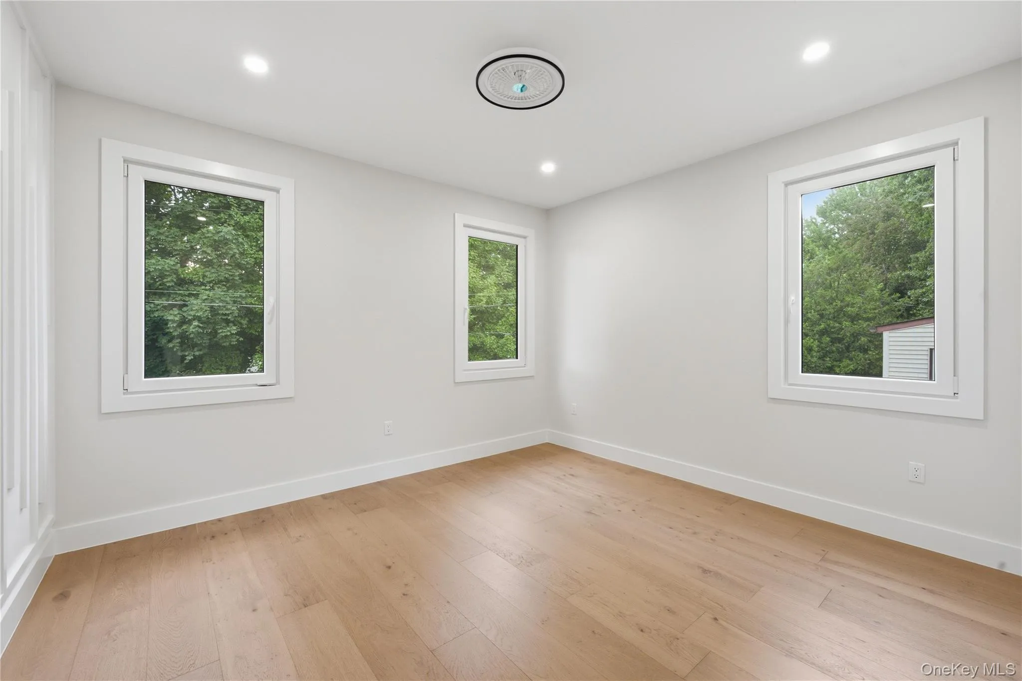 Bright room featuring light wood-finish flooring, crisp white walls, three windows with white trim, recessed ceiling lighting, and a modern flush-mount ceiling fixture Bright room featuring light wood-finish flooring, crisp white walls, three windows with white trim, recessed ceiling lighting, and a modern flush-mount ceiling fixture